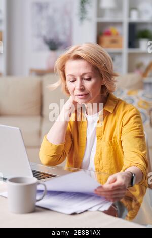 Donna d'affari bionda matura in abbigliamento casualwear intelligente che guarda attraverso i documenti mentre si siede sul posto di lavoro di fronte al notebook in ufficio a casa Foto Stock