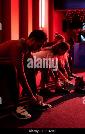 Fila di giovani amici informali seduti su panchina e indossando scarpe da bowling mentre si va a giocare nel centro ricreativo Foto Stock