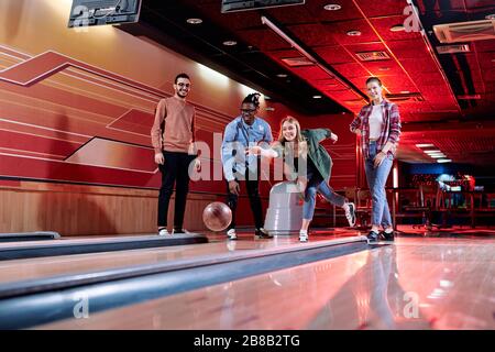 Bella giovane bionda femmina che lancia palla da bowling in pista mentre si trova tra i suoi amici interculturali durante il gioco Foto Stock