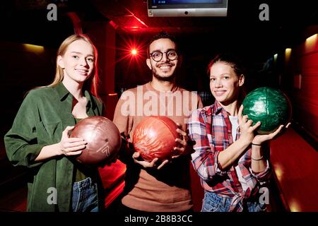 Tre giovani giocatori di bowling interculturali che tengono palle mentre si trovano in una stanza buia sullo sfondo dei vicoli Foto Stock