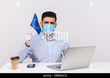 Ufficiale in maschera igienica e guanti in possesso della bandiera UE, preoccupata, avvertimento di epidemia di coronavirus in Europa, quarantena e misure preventive f Foto Stock