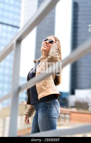 Giovane donna felice in grande città con alti edifici e grattacieli. Donna elegante e sicura con occhiali da sole. Inquadratura verticale. Foto Stock