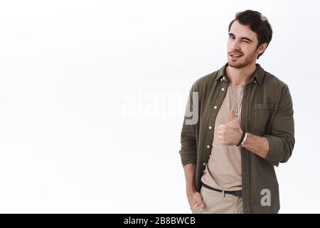 Bello elegante giovane barbuto uomo mostrare thumb-up e wink incoraggiante, lode lavoro impressionante, dire buon lavoro, ben fatto, come qualcosa e raccomandare acquistare Foto Stock