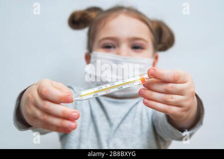 Una bambina in una maschera medica mostra il termometro. Concetto medico di periodo epidemiologico, malattie respiratorie e temperatura elevata Foto Stock