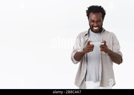 Allegro ragazzo afro-americano bearded che gioca fantastico nuovo gioco, tenendo lo smartphone orizzontalmente in entrambe le mani, sorridendo e guardare lo schermo del telefono cellulare come Foto Stock