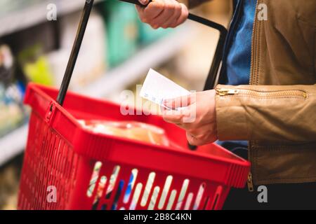 Tenere a mano la lista degli acquisti e il cestino nella corsia del negozio di alimentari. Donna lettura carta, mensola in background. Signora acquistare generi alimentari in supermercato. Foto Stock