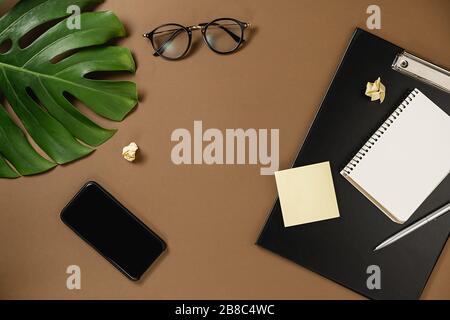Spazio di lavoro con appunti mockup, penna, smartphone, foglie di mostera, occhiali e notebook su sfondo marrone stile minimal. Disposizione piatta, vista dall'alto offic Foto Stock