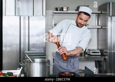 Chef caucasico concentrato che indossa uniforme sta facendo il suo lavoro. Aggiunge pepe alle verdure cotte. Vista frontale. Concetto di cucina Foto Stock