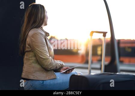 Signora seduta e in attesa di treno in stazione. Donna in piattaforma al tramonto con valigia, biglietto e passaporto. Foto Stock