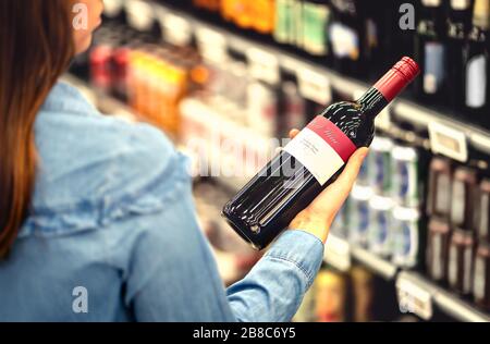 Donna che legge l'etichetta della bottiglia di vino rosso in negozio di liquori o nella sezione di alcool del supermercato. Ripiano pieno di bevande alcoliche. Foto Stock