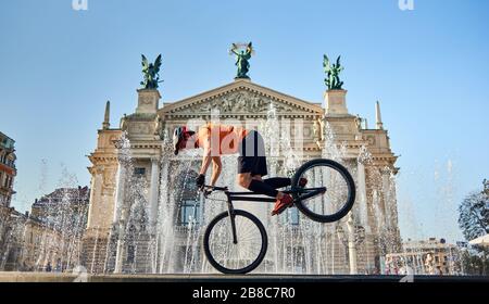 Vista frontale del ciclista sulla ruota anteriore della mountain bike e guardando verso il basso. Il giovane atleta sta pedalando in centro vicino alla fontana. Concetto di attivo. Foto Stock