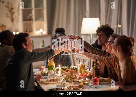 Gruppo di giovani amici felici che brineggiano con il vino e festeggiano la vacanza insieme al tavolo con cena di vacanza a casa Foto Stock
