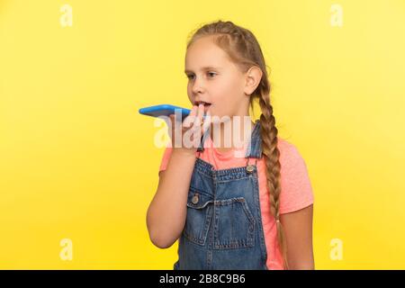 Assistente virtuale. Ritratto di bambina con treccia in tuta denim che parla con il telefono cellulare utilizzando l'applicazione vocale digitale, registrazione audio messa Foto Stock