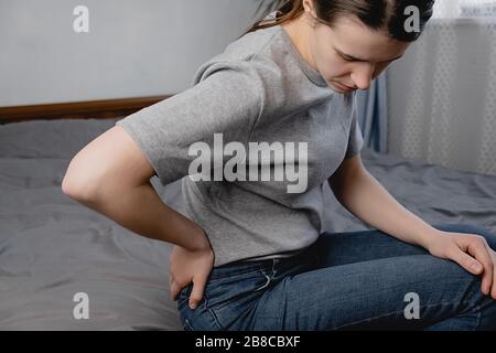 Giovane donna sconvolta che soffre di mal di schiena dopo il sonno, strofinando i muscoli rigidi, ragazza infelice seduta sul letto in camera, sensazione di disagio, a causa di male Foto Stock