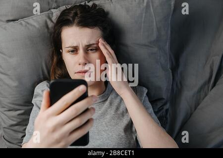 Vista dall'alto della giovane donna scioccata legge notizie terrificanti la mattina sdraiati sul cuscino a letto guardando, fissa il dispositivo smartphone, stupida da rev scioccante Foto Stock