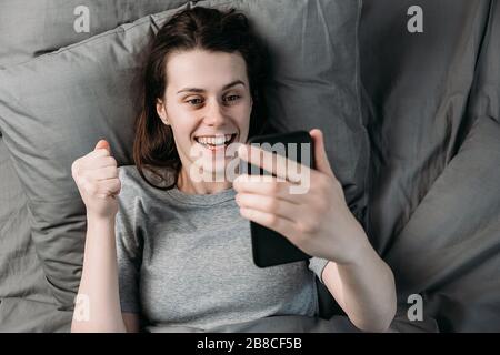 Vista dall'alto della giovane donna entusiasta risvegliata si trova sul letto tenendo in mano lo smartphone urlare con gioia ha ricevuto grandi notizie nel giorno di inizio di mattina. Concetto Foto Stock