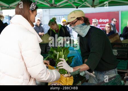 Londra, Regno Unito. 21 marzo 2020. A Venn Street a Clapham gli acquirenti apprezzano la frutta fresca e le verdure disponibili presso il mercato settimanale del sabato. Questo venditore di verdure ha coperto il suo viso con una sciarpa, ma pochissimi indossavano maschere facciali o osservato consiglio di distanziamento sociale. Credit: Anna Watson/Alamy Live News Foto Stock