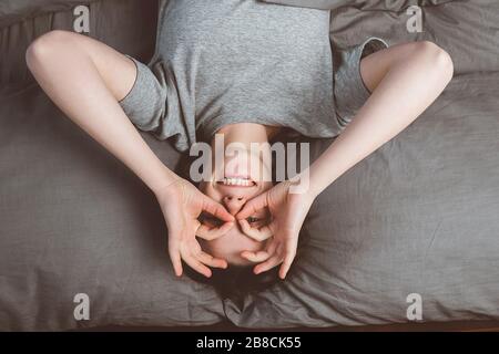 Vista dall'alto di una donna allegra che fa occhiali con le mani, sdraiata in un comodo letto, guardando la macchina fotografica, sorridente giovane donna che indossa una t-shirt grigia Foto Stock