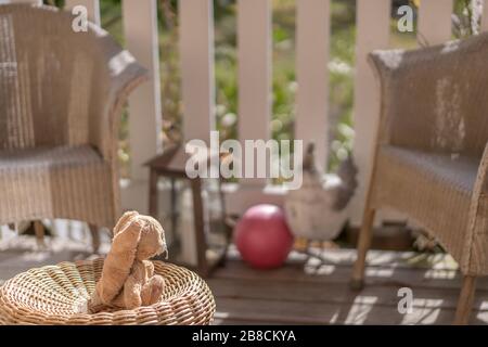 Giocattolo ripieno coniglio pasquale seduta su una sedia di vimini sulla veranda al sole, guardando la palla rosa. Vista ad alta angolazione, primo piano, concetto. Foto Stock