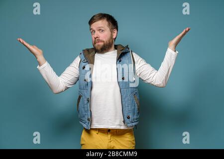 L'uomo barbuto caucasico sembra colpevole, sparge le braccia ai lati e si scusa, su uno sfondo blu. Foto Stock
