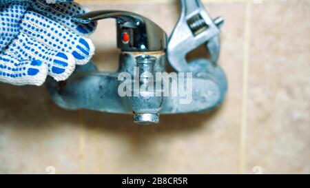 Le mani con i guanti ravvicinati dell'idraulico disinstallano il rubinetto. Il vecchio rubinetto perde e non funziona Foto Stock