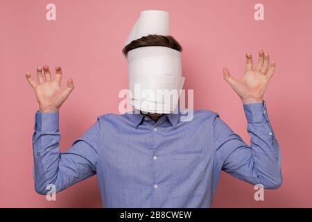 Uomo in camicia blu avvolto il suo viso in una carta come maschera medica cercando di proteggersi dal coronavirus. Studio girato su parete rosa. Foto Stock