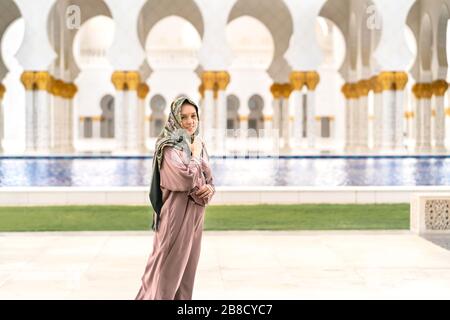 Felice turista donna che indossa sciarpa e abiti nella Grande Moschea Sheikh Zayed di Abu Dhabi. Donna sorridente che cammina in un bellissimo edificio con acqua. Foto Stock