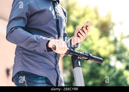 Scooter elettrico e telefono cellulare. Uomo d'affari che utilizza il servizio di noleggio veicoli con smartphone in città strada e parco. Giovane ragazzo millenario. Foto Stock