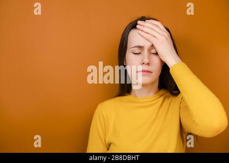 Colpo orizzontale della giovane donna depressa che tiene la mano sulla fronte che è depresso rammaricandosi che cosa ha fatto avere mal di testa, indossa il maglione giallo Foto Stock