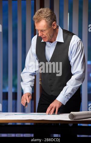 Ingegnere, architetto o uomo d'affari maturo che si trova a una scrivania in un ufficio a tarda notte lavorando con un piano Foto Stock