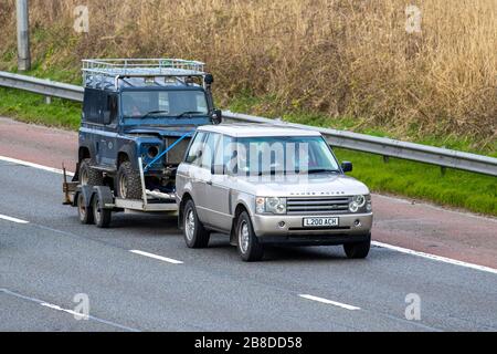 Decrepito di traino di Range Rover ha indebolito il vecchio Land Rover Defender. Un progetto di restauro, il fienile trainato trova; traffico veicolare del Regno Unito, trasporto, veicoli in movimento, veicolo di restauro, strade, motori di traino, rimorchio per auto sulla autostrada M6 Foto Stock