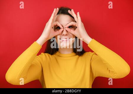 Allegra giovane donna divertente che si diverte a fare gli occhiali forma con le mani, fa faccia divertente con gli occhi spalancati, vestita in maglione giallo Foto Stock