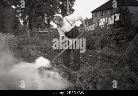 1953, storico, al di fuori di un cottage rurale, un uomo anziano, sigaretta in bocca e indossare pantaloni e bretelle di pinstrip e rastrello in mano, lavorando nel suo giardino nel patch di verdure bruciare vecchie foglie su un falò, Inghilterra, Regno Unito. Foto Stock