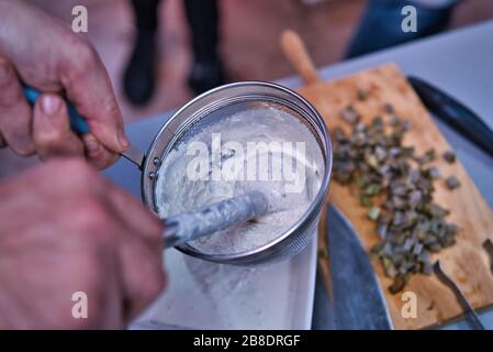 Primo piano di un setaccio da cucina che versa la crema in una ciotola bianca. Cucina fatta in casa e naturale. Cuoco informale. Pentole vegetali verdi fuori di fuoco su legno Foto Stock