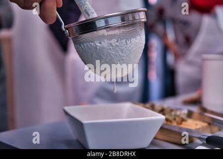 Primo piano di un setaccio da cucina che versa la crema in una ciotola bianca. Cucina fatta in casa e naturale. Cuoco informale Foto Stock