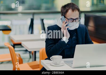 Un lavoratore uomo molto impegnato lavora in quarantena, legge le informazioni online sul computer portatile, indossa una maschera medica protettiva, chiama qualcuno tramite cellulare, c Foto Stock