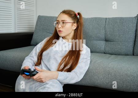 Una ragazza dai capelli rossi con occhiali, con lunghe code, si siede sul pavimento in un mezzo giro appoggiandosi su un divano con una console di gioco Foto Stock