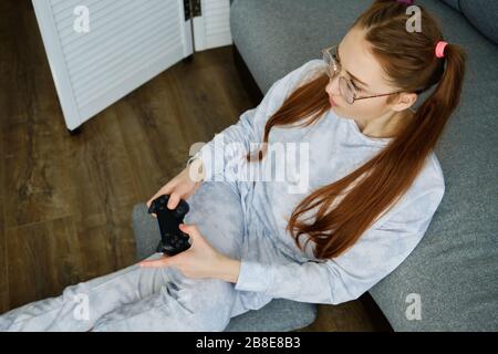 Ragazza rossa con occhiali, con lunghe code si siede sul pavimento in un mezzo giro appoggiato su un divano con una console di gioco, girato dall'alto Foto Stock