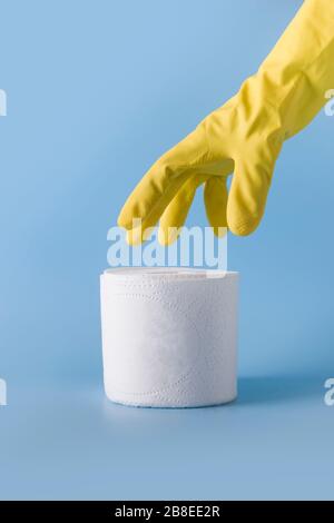 La mano con guanti gialli raggiunge il rotolo di carta igienica bianca su un tavolo blu. Inquadratura verticale. Concetto di carenza di materie prime essenziali nel Coronavirus t Foto Stock
