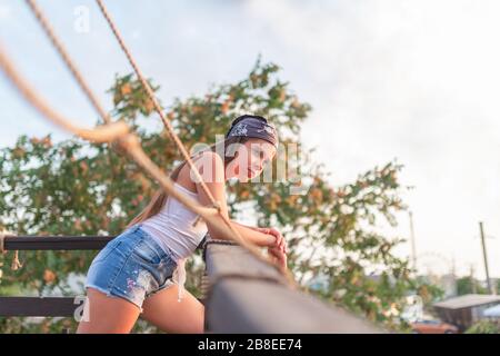 Vista laterale di una ragazza carina adolescente in pantaloncini e un cappello si erge sul balcone aperto stilizzato come una nave pirata la calda serata estiva contro un cielo blu. C Foto Stock