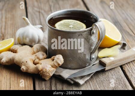 Tazza di tè allo zenzero con limone, radici di zenzero, aglio e coltello sul tavolo da cucina in legno. Foto Stock