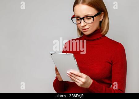 Ritratto aziendale di donna elegante e sicura con dolcevita rosso, indossando occhiali ottici, tenendo e digitando sul computer tablet, isolato su ba grigia Foto Stock