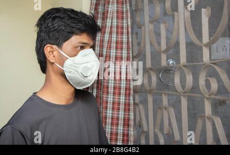 Giovane uomo con maschera medica che guarda attraverso la finestra - concetto di isolamento domestico o di quarantena a causa di 19 covid o scoppio di coronavirus Foto Stock