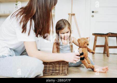 Cute bambino ragazza lungo capelli lisci grandi occhi grigi indossare pigiama gioco di orso giocattolo con la madre in camera rimanere a casa Foto Stock