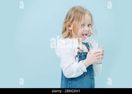 Perky bambina in tute jeans sorseggiando milkshake attraverso una paglia su blu Foto Stock