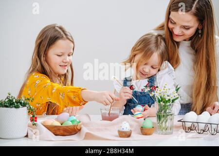 Giovane madre seduta con le sue due figlie dipingere le uova di pasqua dietro un tavolo Foto Stock