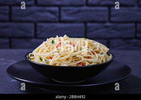 Pasta Aglio, olio e Peperoncino, spaghetti italiani con aglio, peperoncino e olio d'oliva in una ciotola nera su un tavolo di cemento con parete in mattoni a fondo Foto Stock