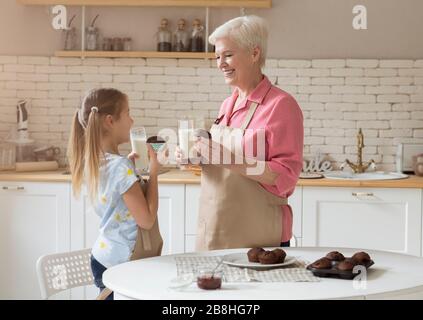 Adorabile ragazza con la nonna mangiare muffin appena sfornati e bere latte in cucina Foto Stock