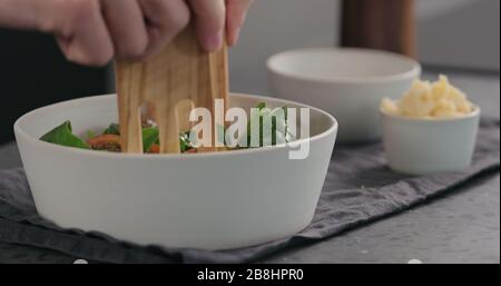 uomo che mescola insalata con pomodori kumato e spinaci in ciotola bianca witj server di legno di oliva, foto ampia Foto Stock