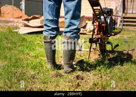 Coltivatore in stivali di gomma e jeans blu arows un terreno con trattore a motore di smal. Foto Stock